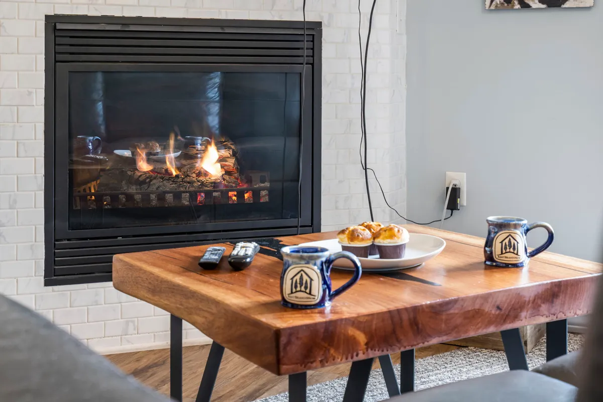 A cozy sitting area with a seasonal propane fireplace, a wooden coffee table set with mugs and plated food, and comfortable seating—conveying warmth and intimacy. The fire glows softly, enhancing the romantic, relaxing atmosphere.