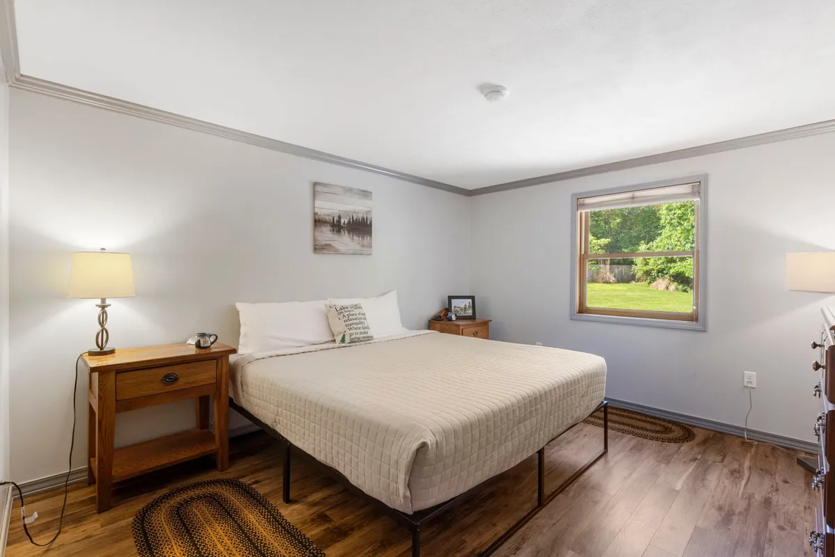 A serene bedroom scene featuring a plush bed with soft linens, a large window allowing natural light to stream in, a bedside table with a lamp, and a piece of calming artwork above the bed. The overall atmosphere is peaceful and perfect for unwinding.