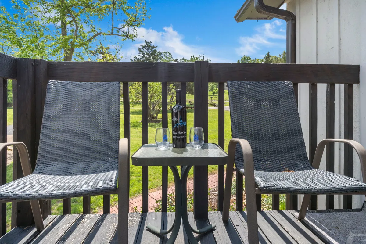 The image showcases a charming outdoor deck featuring two inviting chairs and a small table set with a bottle of wine and glasses. The deck overlooks a lush green lawn, and the soft glow of evening sunlight promises serene sunset views, perfect for unwinding or sharing a quiet moment.