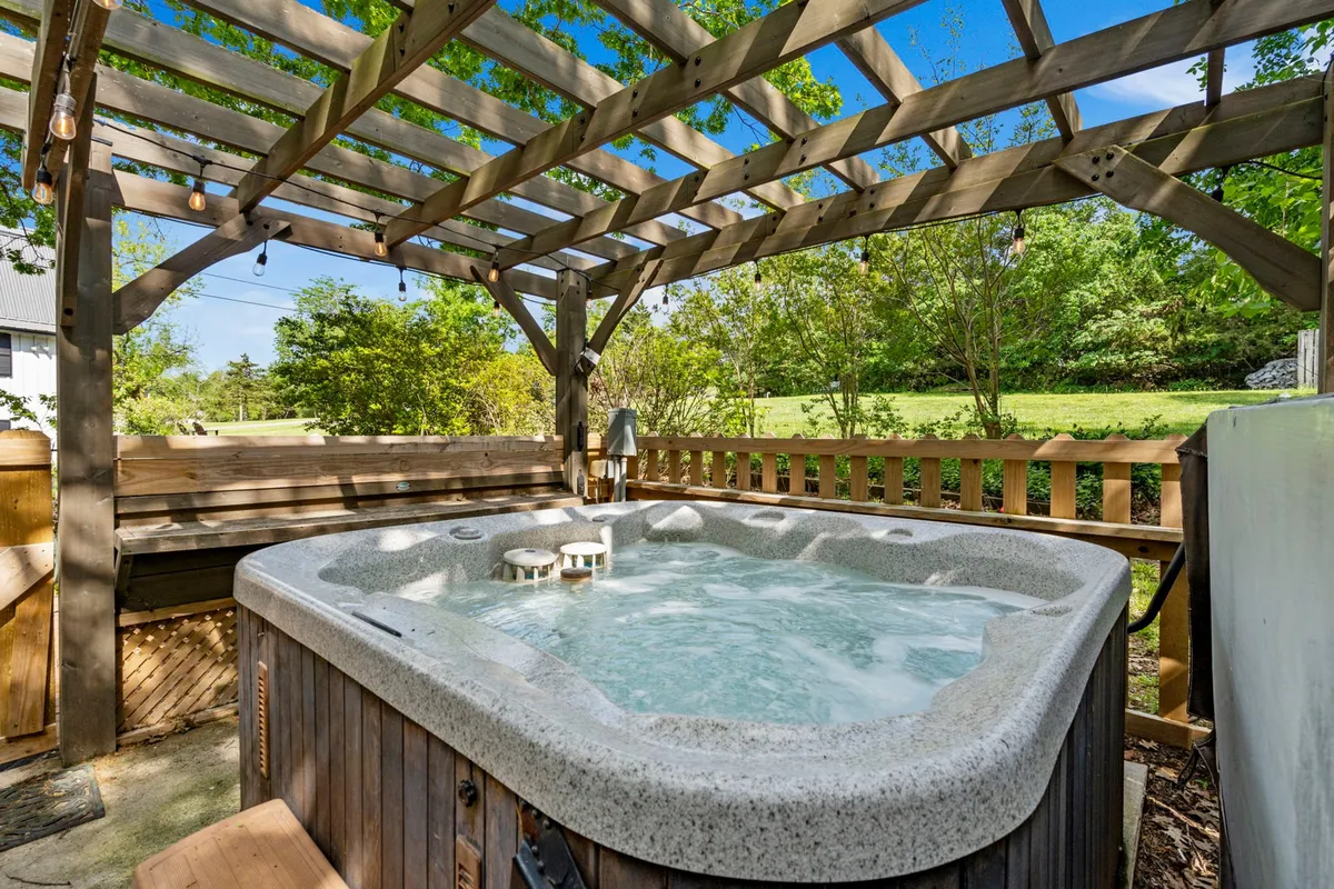 An inviting outdoor hot tub sits beneath a wooden pergola, surrounded by lush greenery. The setting is tranquil yet social, offering a comfortable space for guests to relax, socialize, and enjoy nature. The hot tub is the focal point, hinting at shared experiences and relaxation.