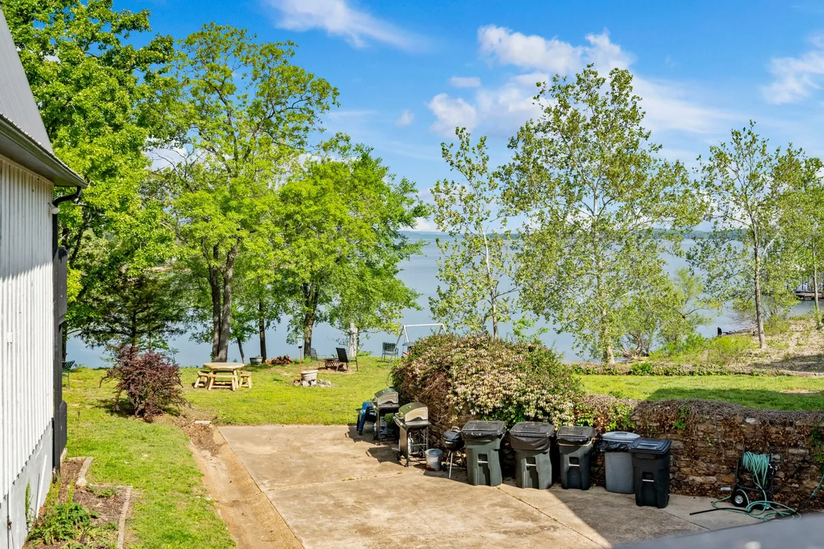 The image shows a tranquil waterfront view with lush greenery and a welcoming patio area, perfect for picnicking or relaxing outdoors. The lake and surrounding nature create an inviting environment for gatherings and recreation.