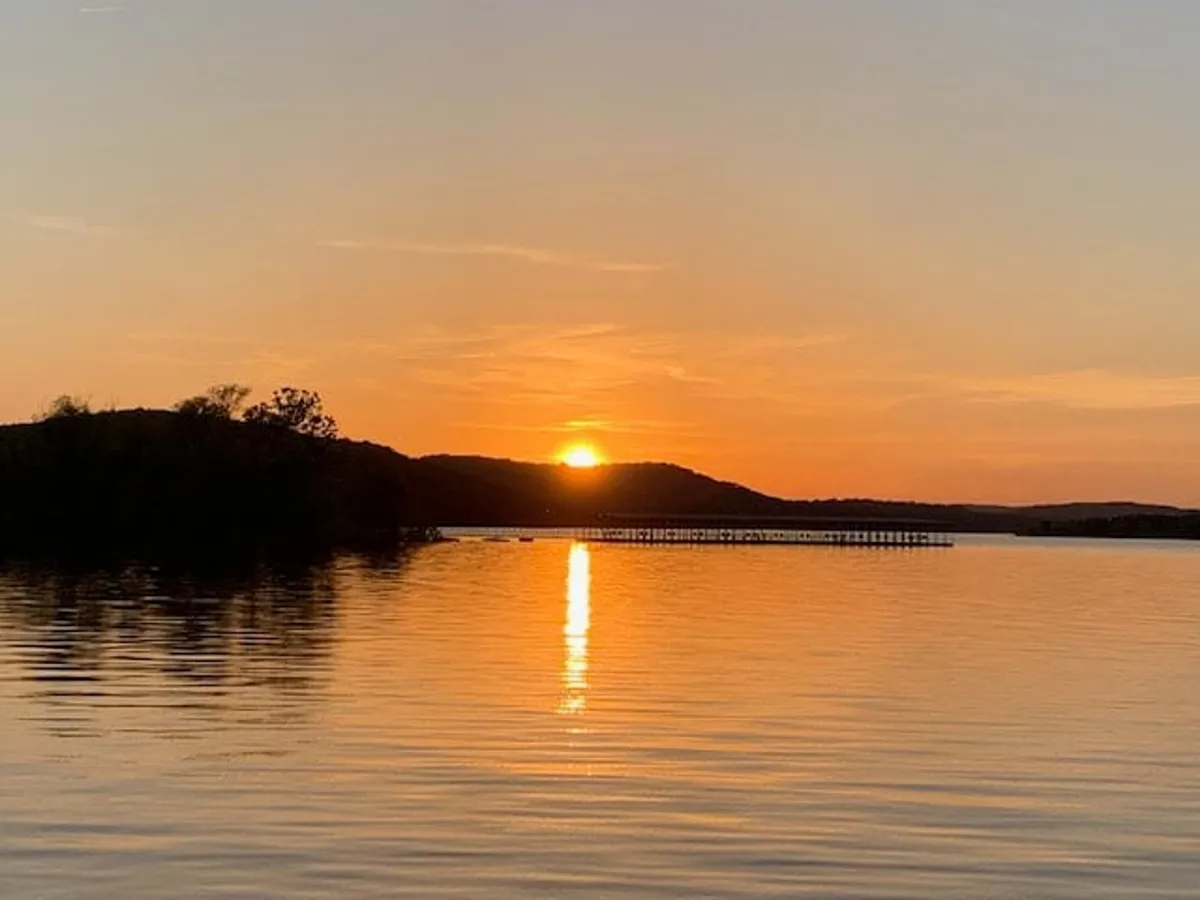 This image captures a breathtaking sunset over Table Rock Lake, with the water reflecting warm hues and a distant pier stretching out towards gently rolling hills. The serene atmosphere evokes romance and tranquility, perfectly aligning with the idea of couples savoring the evening together by the water.